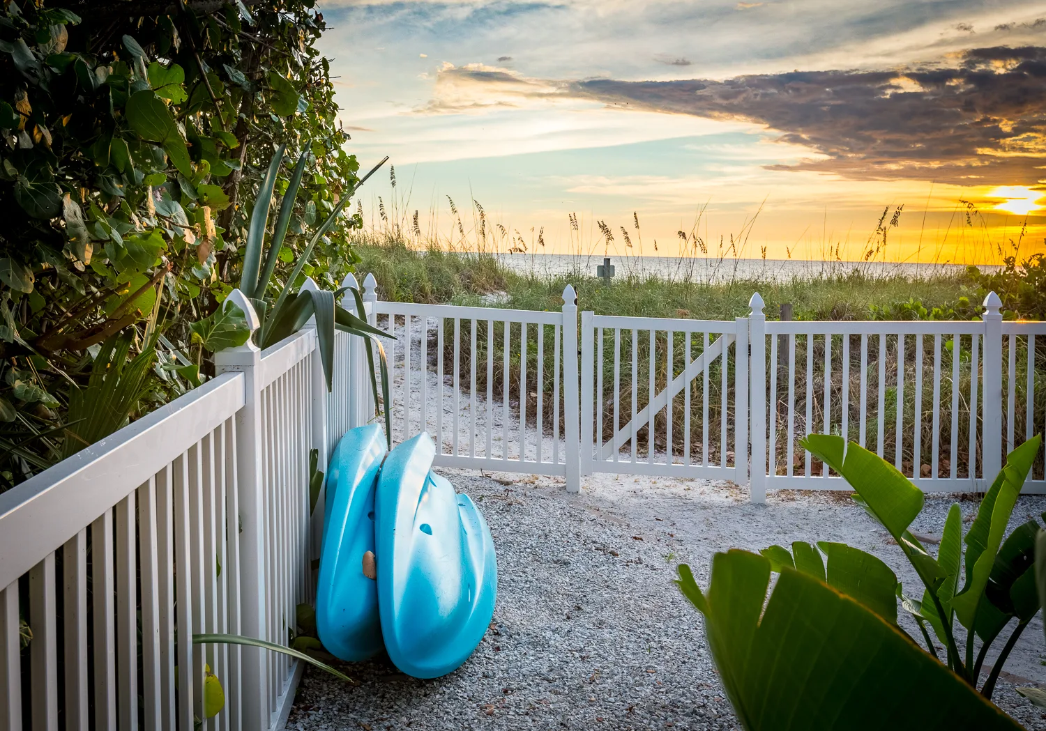 Two blue kayaks by a white gate on the sandy beach path at sunset