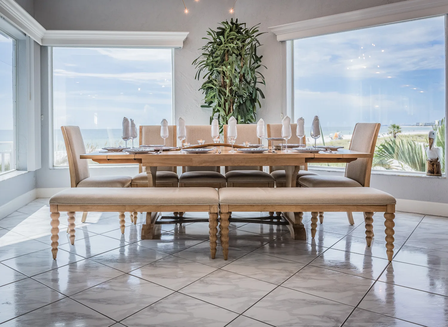 Set dining table with upholstered chairs and benches beside beach-view windows