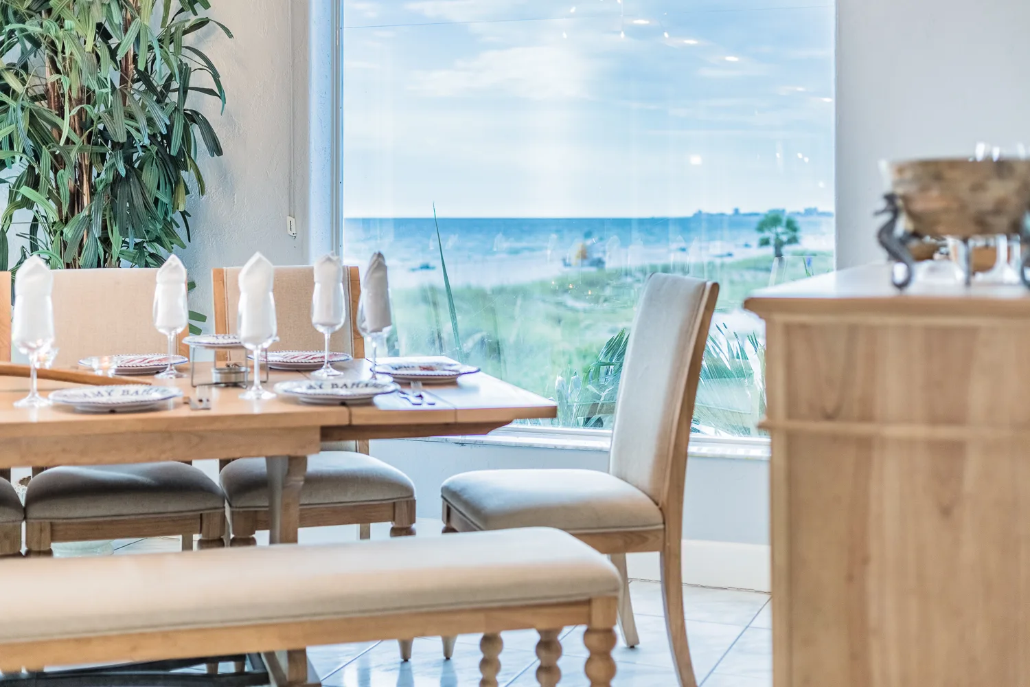 Dining table set with folded napkins beside a window overlooking the beach