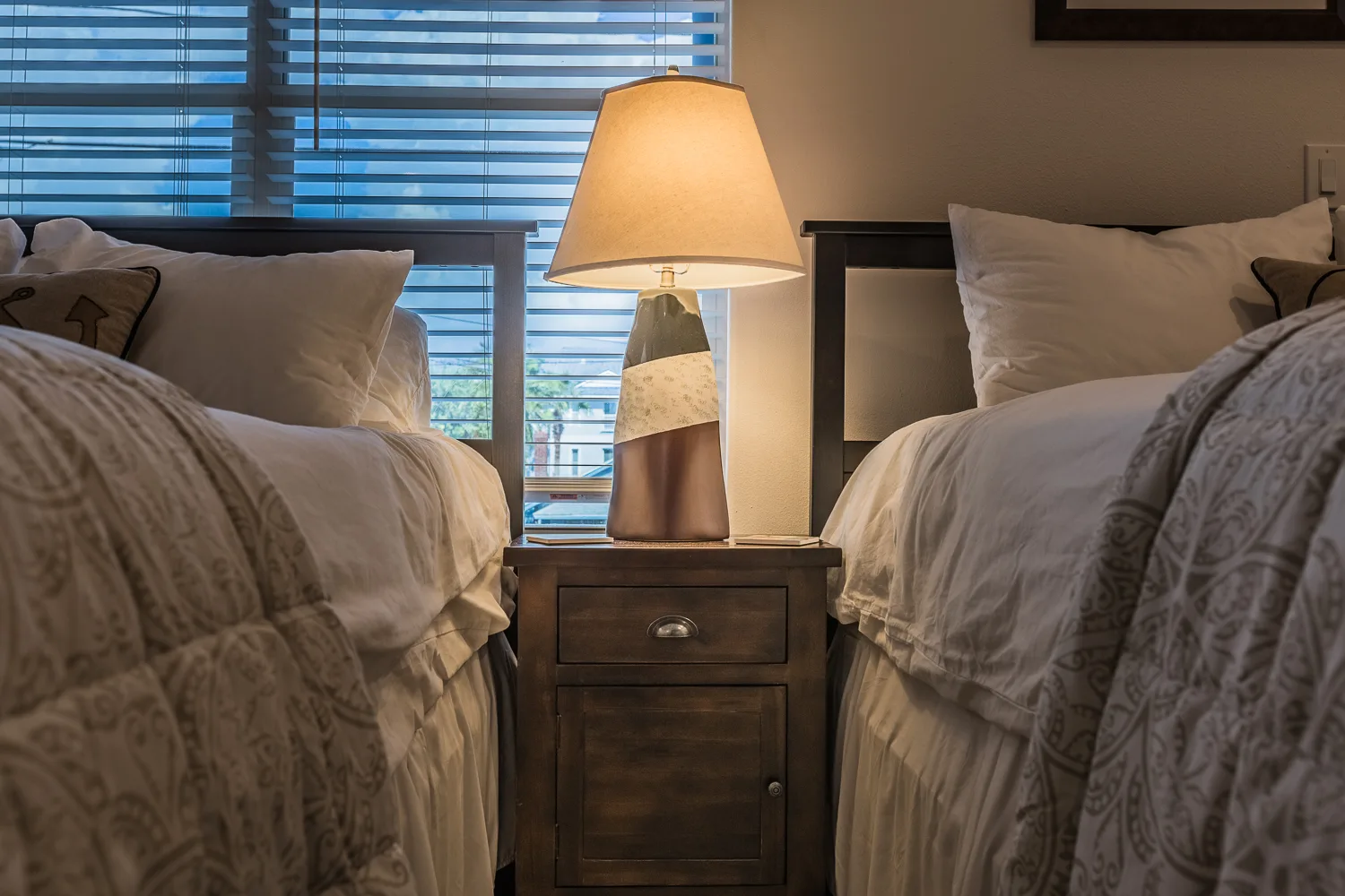 Lamp glowing on a nightstand between two beds at a shuttered window
