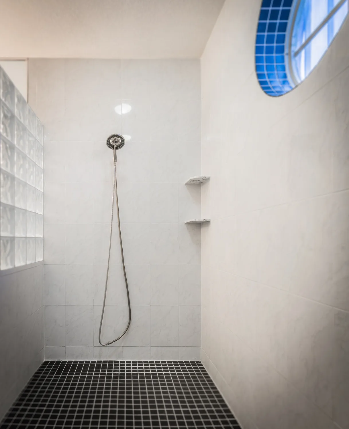 Walk-in shower with handheld fixture, glass block wall and oval blue-tiled window