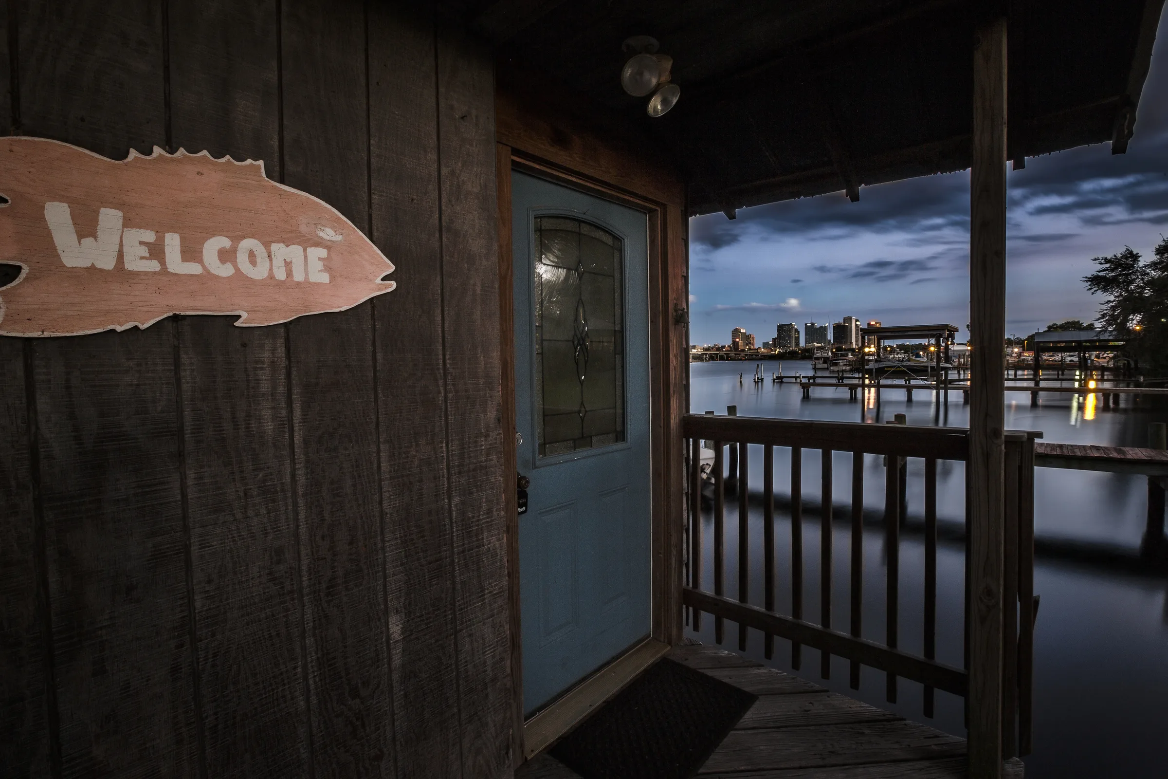 Fish-shaped welcome sign beside blue door on rustic waterfront porch at dusk