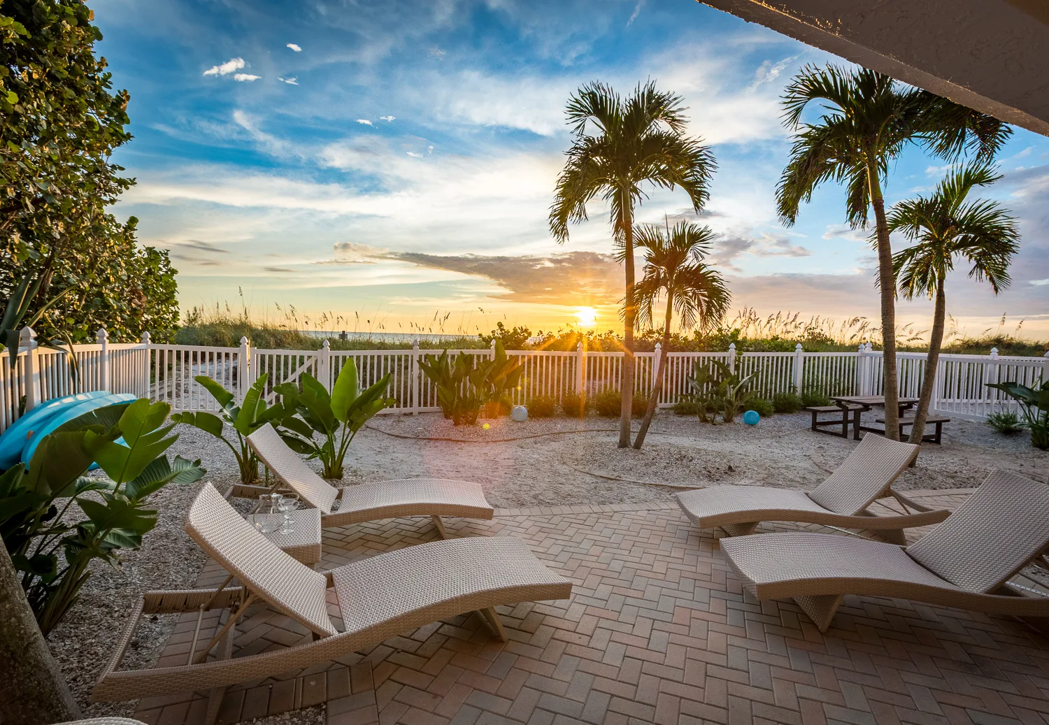 Sunset over the dunes behind lounge chairs, palms and a white fence
