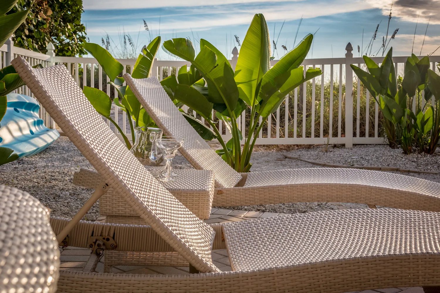Wicker lounge chairs among tropical plants inside a white beachfront fence