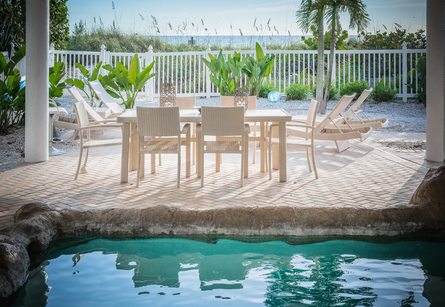 Covered poolside dining table with beach views beyond a white fence