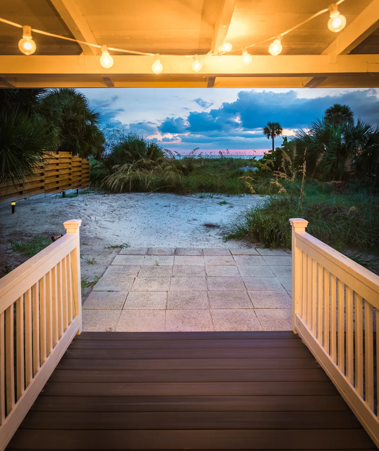 Deck with string lights leading to a sandy beach path at dusk