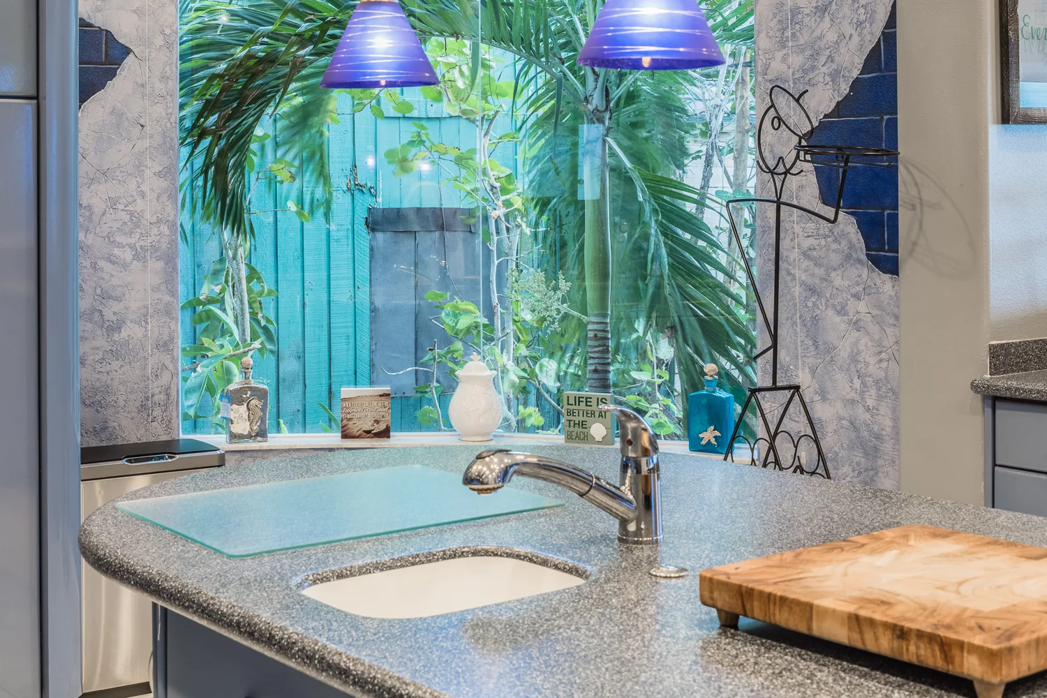 Kitchen sink beneath purple pendants with lush tropical window view
